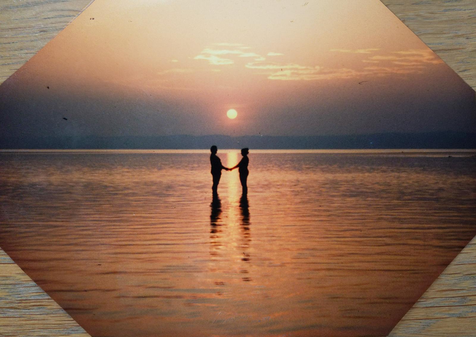 Two people standing together holding hands, silhouetted against a sunset over still water