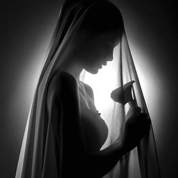 Black-and-white photograph of a veiled figure bowed over a calla lily, lit from behind